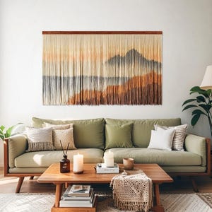 May include: A large rectangular wall hanging made of vertical fiber strands suspended from a wooden rod. The fibers are dyed in a gradient pattern featuring shades of cream, tan, orange, and dark gray, creating a landscape-like mountain scene.