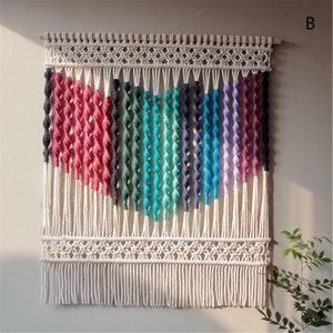 May include: A macrame wall hanging with a rainbow gradient design. The piece features a wooden dowel at the top, intricate knotwork, and a fringe at the bottom. The colors include pink, black, green, teal, blue, and purple.