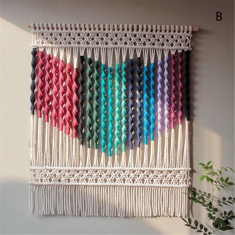 May include: A macrame wall hanging with a rainbow gradient design. The piece features a wooden dowel at the top, intricate knotwork, and a fringe at the bottom. The colors include pink, black, green, teal, blue, and purple.