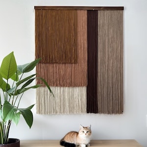 May include: A woven wall hanging with a wooden frame. The textile art features a layered design with shades of brown, tan, and cream. A houseplant and a cat are in the foreground. The cat is brown, tan, and black.