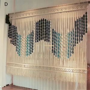 May include: A large, off-white macrame wall hanging with a geometric design. The design incorporates twisted rope elements in shades of blue and white, creating a wave-like pattern. The piece is finished with a fringed bottom and decorative top.