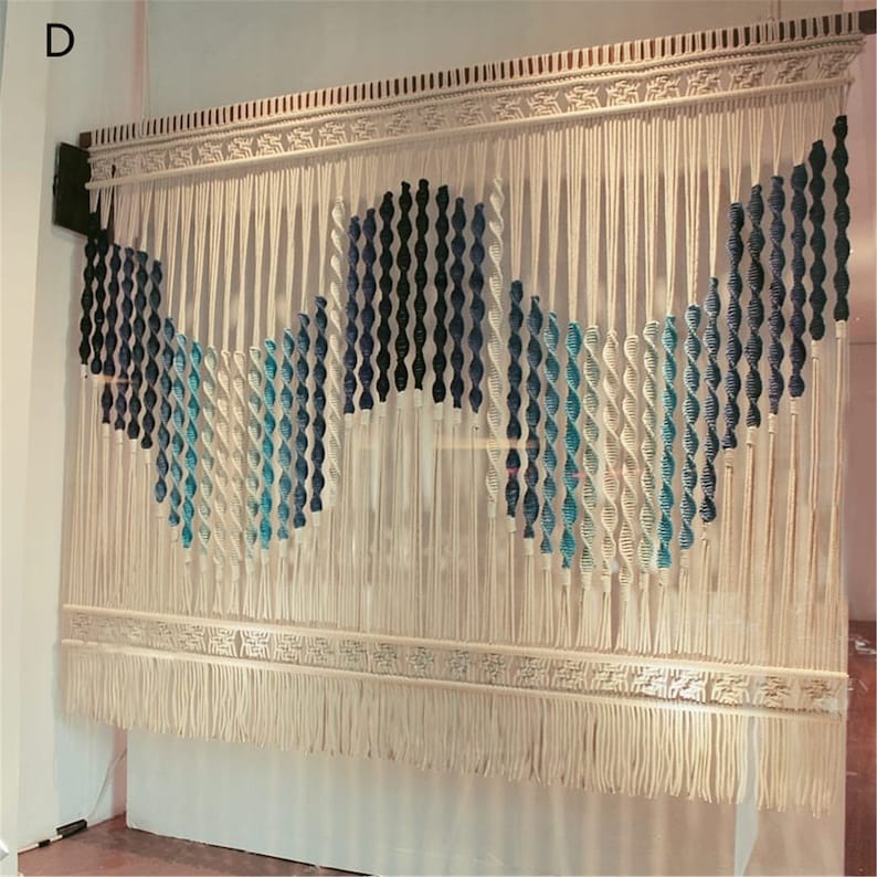 May include: A large, off-white macrame wall hanging with a geometric design. The design incorporates twisted rope elements in shades of blue and white, creating a wave-like pattern. The piece is finished with a fringed bottom and decorative top.