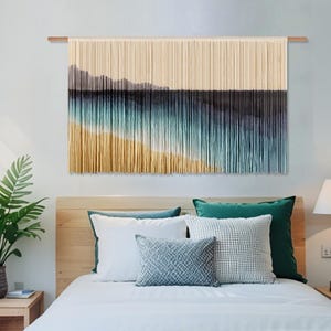 Puede incluir: Un tapiz de pared tejido que representa una escena costera. La obra de arte presenta un degradado de colores, que va del beige al amarillo, turquesa y azul oscuro, sugiriendo una playa y el océano. Está montado en una barra de madera.