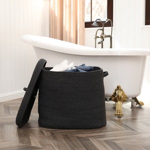 May include: A dark gray woven laundry basket with a matching lid, partially open, revealing folded clothes. The basket is in a bathroom setting, near a white bathtub with gold accents.
