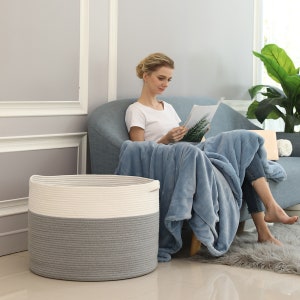 May include: A large gray and white rope storage basket with a handle. The basket is sitting on a light gray floor. A woman is sitting on a couch in the background.