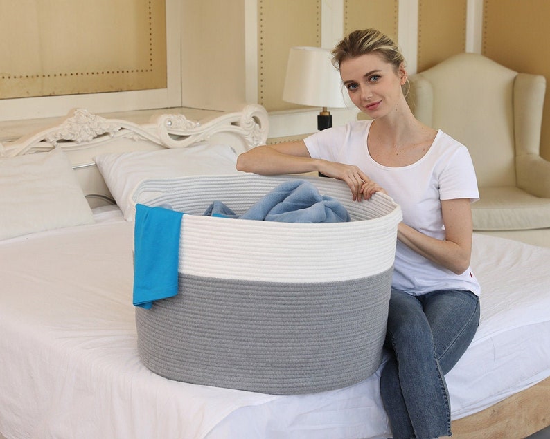 May include: A large, gray and white rope storage basket with a blue towel and a gray blanket inside. The basket is sitting on a white bed.
