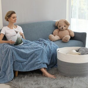 May include: A woman sits on a blue couch with a plush blue throw blanket. A teddy bear sits on the couch next to her. A white and gray rope storage basket sits on the floor in front of the couch.