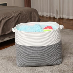 May include: A white and gray rope storage basket with a blue and white blanket and a red and green toy inside.