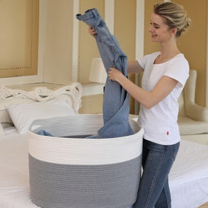 May include: A person is putting a pair of blue jeans into a large, gray and white rope storage basket. The basket is on a white bed.