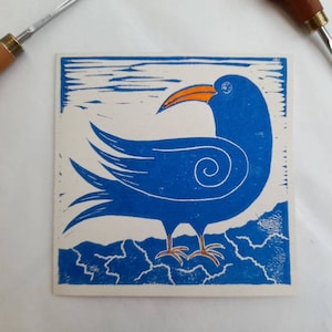May include: A blue and white linocut print of a stylized bird with an orange beak and feet. The bird is depicted on a square, with a wavy design at the bottom and a horizon line at the top. Carving tools are in the background.