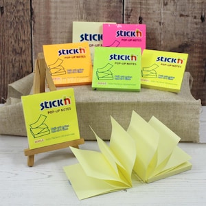Puede incluir: Una colección de notas adhesivas Stick'n Pop-Up en varios colores: amarillo, naranja, verde y rosa. Las notas, de 76 mm x 76 mm, se exhiben sobre un caballete de madera y una superficie de arpillera. El texto 'Pull out notes one at a time' es visible.