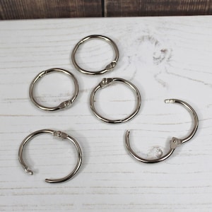 May include: Five silver binder rings are displayed on a white wooden surface. The rings are in various states of closure, demonstrating their use. The shiny metal rings are designed for organizing documents or other items.