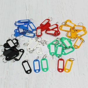 May include: A collection of rectangular plastic key tags with metal split rings, featuring white paper inserts for labelling. The tags are available in a variety of colours including blue, red, yellow, green, white, and black, arranged in a scattered group.