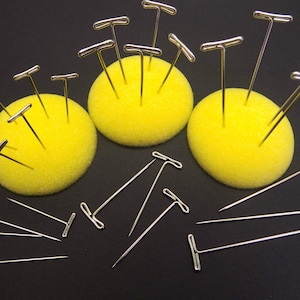 Könnte beinhalten: Drei gelbe Schaumstoff-Stecknadelkissen mit mehreren silbernen T-Nadeln, die in sie gesteckt sind. Die Stecknadelkissen befinden sich auf einer schwarzen Oberfläche.
