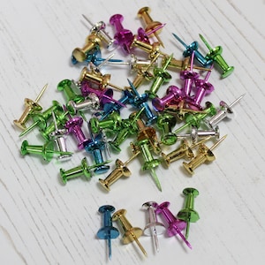 May include: A large assortment of colourful push pins in various metallic shades, including gold, silver, pink, green, and blue. The push pins are scattered on a white wooden surface.