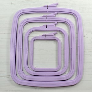 May include: Four lavender-coloured square embroidery hoops of different sizes are nested together. Each hoop has a tightening screw and is labelled with a number and millimetre measurement. The largest hoop is 250 mm, and the smallest is 95 mm.