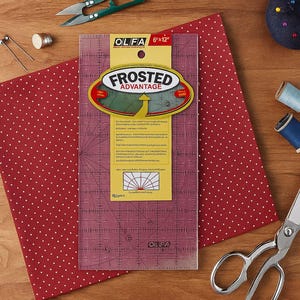 Può includere: Un righello da quilting OLFA trasparente e smerigliato da 15,24 cm x 30,48 cm con linee a griglia e la scritta "FROSTED ADVANTAGE" su un'etichetta gialla. Il righello è su un tessuto rosso a pois, con strumenti di cucito sullo sfondo.