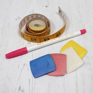 May include: A white fabric marking pen, a yellow tape measure, and four fabric marking tools in blue, pink, white, and yellow. The tape measure reads "Adorn Choose Are True".