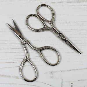 May include: Two pairs of silver-toned, decorative scissors with ornate handles. The scissors have a pointed tip and are designed for delicate tasks.