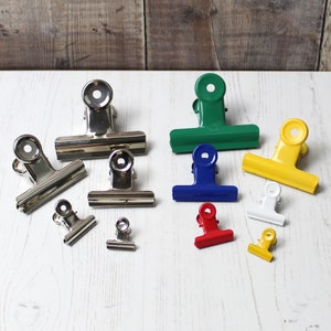 May include: An assortment of metal binder clips in various sizes and colours, including silver, green, blue, yellow, red, and white. The clips are arranged on a white surface.