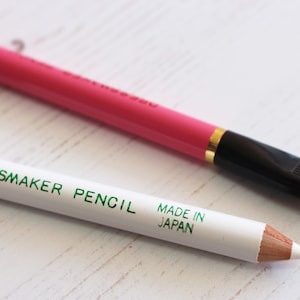 May include: Two dressmaker pencils. One is white with green lettering that says "Dressmaker Pencil Made in Japan". The other is pink with a black brush tip.
