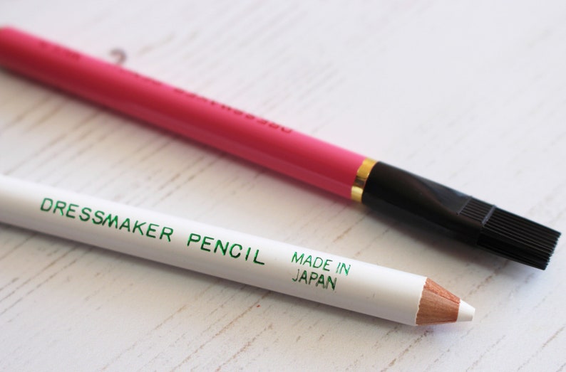 May include: Two dressmaker pencils. One is white with green lettering that says "Dressmaker Pencil Made in Japan". The other is pink with a black brush tip.
