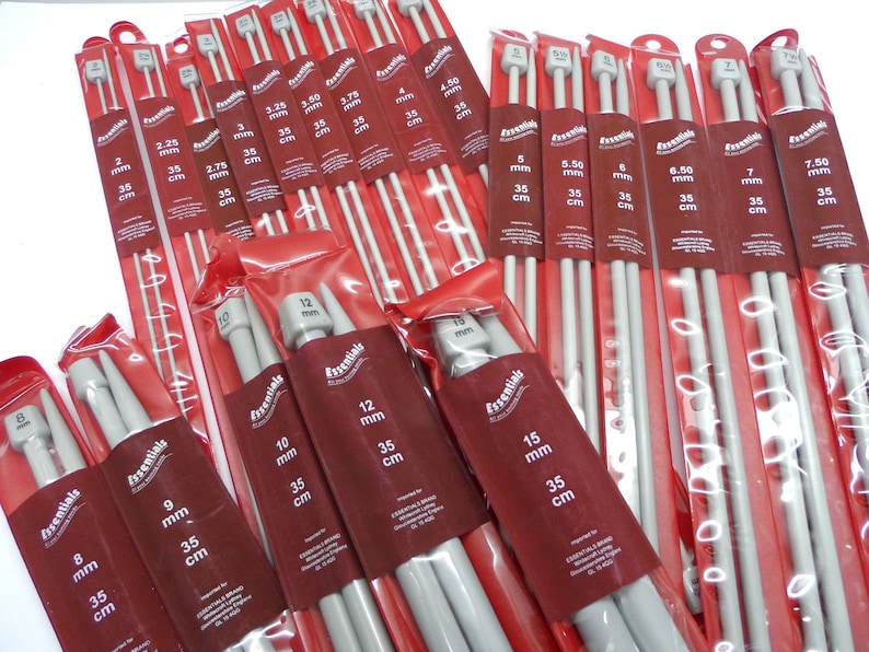 May include: A set of 12 pairs of grey knitting needles in various sizes, ranging from 2 mm to 15 mm. The needles are individually packaged in clear plastic bags with red labels that indicate the size in millimetres and centimetres. The labels also include the word "Essentials".