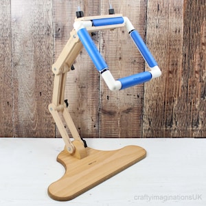Può includere: Un supporto per ricamo in legno con braccio regolabile e telaio rettangolare. Il telaio è composto da angolari in plastica bianca e barre rivestite in schiuma blu. La base è un pezzo di legno piatto a forma di T con il testo Crafty Imaginations.