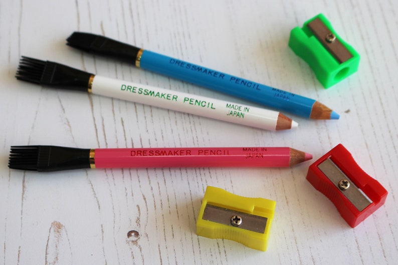 May include: Three dressmaker pencils with built-in brushes, one blue, one white, and one pink, with a yellow, green, and red pencil sharpener.