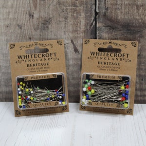 Könnte beinhalten: Zwei Packungen mit Glaskopfnadeln, eine Packung ist 30 mm x 0,60 mm und die andere 48 mm x 0,80 mm. Beide Packungen sind mit "Whitecroft England Heritage Premium Quality" gekennzeichnet. Die Nadeln befinden sich in einem durchsichtigen Plastikbehälter und haben farbige Glasköpfe.