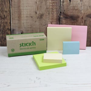 Può includere: Una collezione di post-it in vari colori pastello, tra cui rosa, giallo e blu, accanto a una scatola di "stick'n RECYCLED NOTES". La scatola è marrone con dettagli verdi. Le note sono disposte su una superficie di legno bianco.