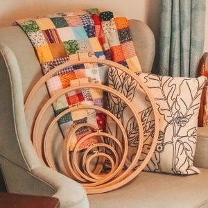 May include: A set of five wooden embroidery hoops of varying sizes, nested together. The hoops are sitting on a light brown armchair with a colorful patchwork quilt and a pillow with a floral design.