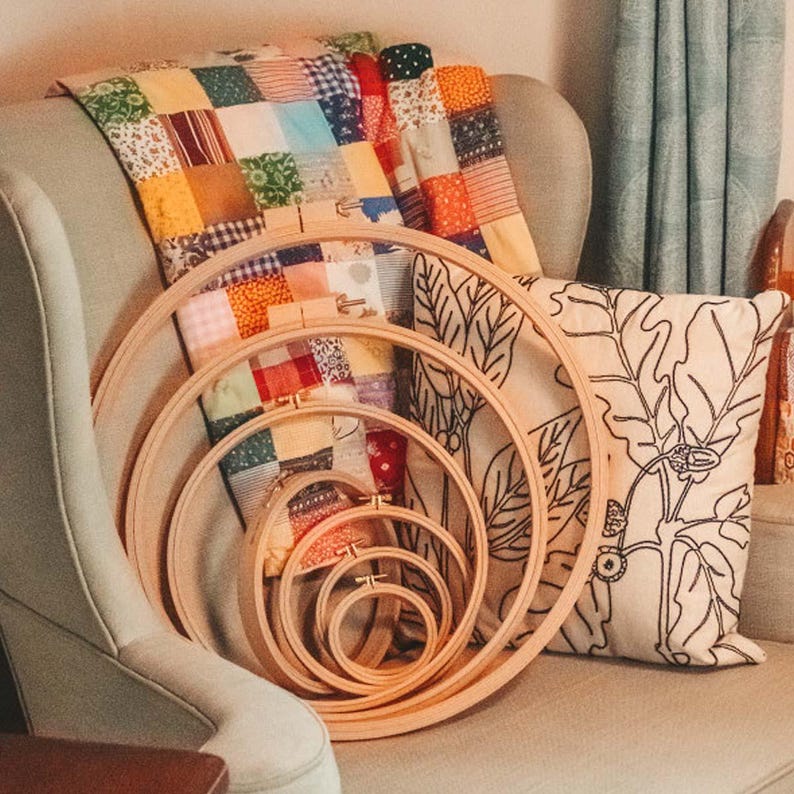 May include: A set of five wooden embroidery hoops of varying sizes, nested together. The hoops are sitting on a light brown armchair with a colorful patchwork quilt and a pillow with a floral design.