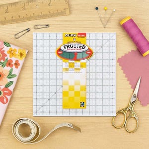 Op de afbeelding: Een overzicht van naai- en quiltbenodigdheden. Inclusief een mat, antislip liniaal, meetlint, roze garenklos, stof, gouden schaar, spelden en een houten liniaal. De liniaal heeft rasterlijnen en de tekst "FROSTED ADVANTAGE".