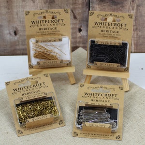 Puede incluir: Cuatro paquetes de alfileres de costura, cada uno con un acabado diferente: dorado, negro, latón y plateado. Los paquetes están etiquetados con "Whitecroft England Heritage" y presentan las palabras "Premium" y "Quality".