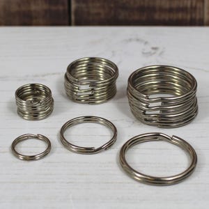 Könnte beinhalten: Eine Ansammlung von silberfarbenen Schlüsselringen in verschiedenen Größen, angeordnet auf einer weißen Holzoberfläche. Die Metallringe sind gestapelt und haben ein geteiltes Design zum Befestigen von Schlüsseln.