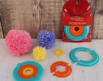 May include: A set of three colorful plastic pom pom makers in blue, orange, and yellow. The set includes two smaller pom pom makers and one larger pom pom maker. The pom pom makers are packaged in a clear plastic bag with a red header that says "WHITECRAFT essentials SPLIT POM POM MAKER www.whitecraft.co.uk".