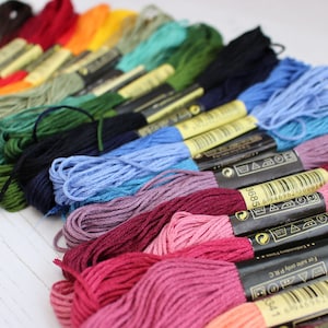 May include: A close-up of a variety of embroidery floss spools in different colors, including red, pink, blue, green, and yellow. The spools are arranged in a row, with the labels facing the viewer. The labels include the brand name, color number, and care instructions.