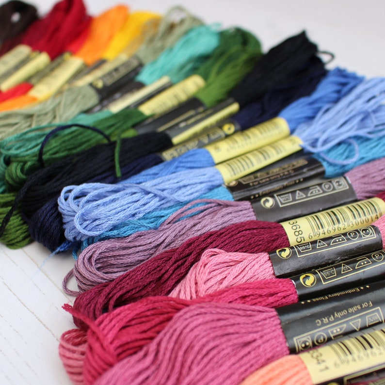 May include: A close-up of a variety of embroidery floss spools in different colors, including red, pink, blue, green, and yellow. The spools are arranged in a row, with the labels facing the viewer. The labels include the brand name, color number, and care instructions.