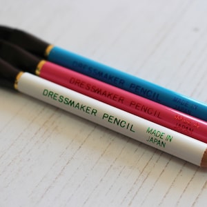 May include: Three dressmaker pencils with black brush tips. The pencils are white, pink, and blue. Each pencil has the text "Dressmaker Pencil" and "Made in Japan" printed on it.