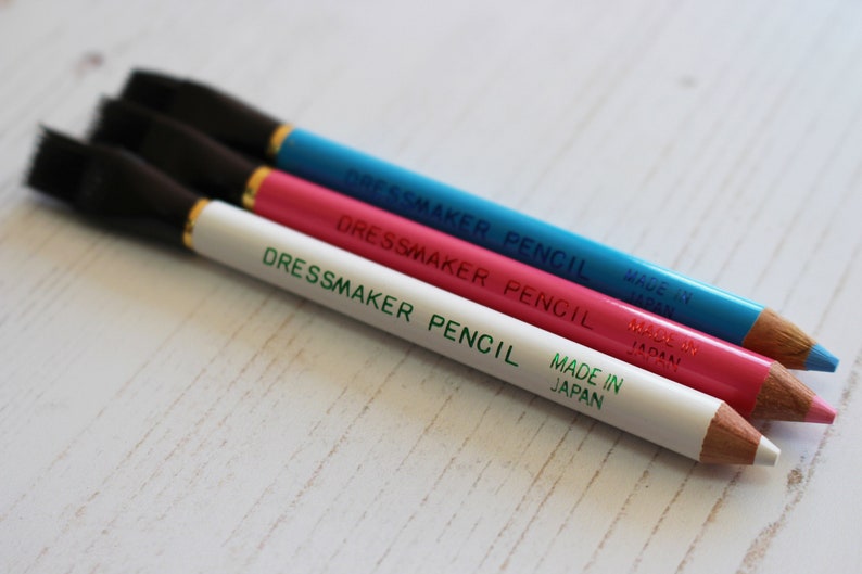 May include: Three dressmaker pencils with black brush tips. The pencils are white, pink, and blue. Each pencil has the text "Dressmaker Pencil" and "Made in Japan" printed on it.