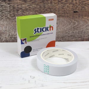 May include: A roll of grey repositionable double-sided tape beside a box. The box is white with green and orange accents and the text "Stick'n". The tape is 2.5cm wide and 12m long.