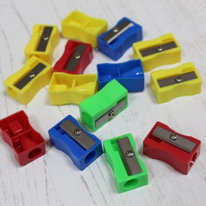 Op de afbeelding: Een verzameling van 11 kleurrijke plastic potloodslijpers. De slijpers zijn in verschillende kleuren, waaronder rood, blauw, groen, geel en wit. Elke slijper heeft een klein metalen mesje om potloden te slijpen.