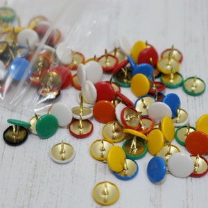 May include: A large assortment of colourful push pins in a plastic bag. The push pins are in various colours, including red, yellow, blue, green, and white. The push pins are a popular office supply used for attaching papers to notice boards.