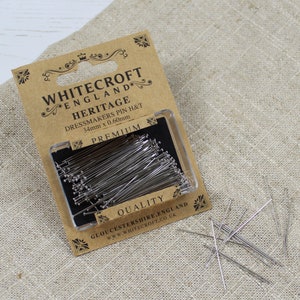 Puede incluir: Un paquete de alfileres de sastre plateados en un recipiente de plástico transparente. El paquete está etiquetado con "Whitecroft England Heritage Dressmakers Pin H&T 34mm x 0.60mm Premium Quality Gloucestershire, England www.whitecroft.co.uk"