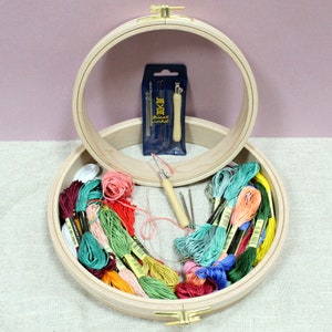 May include: A wooden embroidery hoop containing a selection of colourful embroidery floss spools. The hoop is 10 inches in diameter and has a brass screw closure. The floss is arranged in a rainbow pattern. There is also a small pack of embroidery needles and a wooden needle threader.