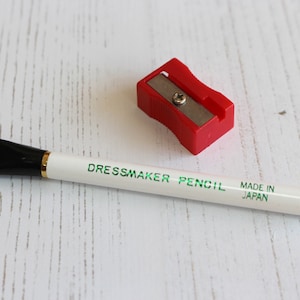May include: White dressmaker pencil with a black brush on one end and a sharpener. The pencil is labeled "Dressmaker Pencil" and "Made in Japan".