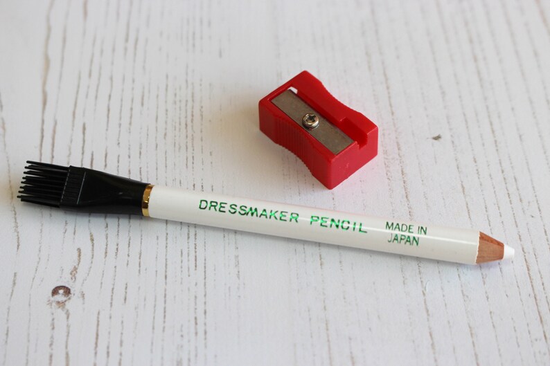 May include: White dressmaker pencil with a black brush on one end and a sharpener. The pencil is labeled "Dressmaker Pencil" and "Made in Japan".
