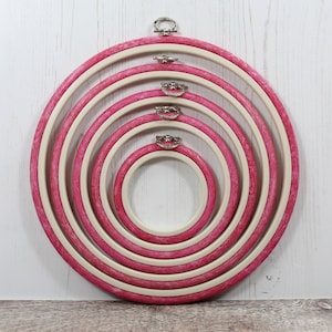 May include: Set of five embroidery hoops with a pink and white wood finish. The hoops are nested inside each other, with the largest hoop at the bottom and the smallest hoop at the top.