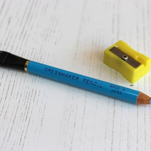 May include: A blue dressmaker pencil with a black brush on one end and the words "Dressmaker Pencil Made in Japan" printed on the side. A yellow pencil sharpener is also pictured.